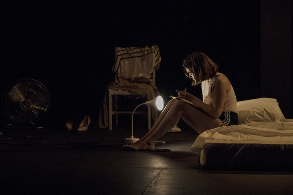 Ambiente semi oscuro, una pequeña lámpara de mesa alumbra a una mujer sentada sobre el costado en un colchón (que está sobre el piso), con los pies en el piso y anotando algo sobre una libreta que apoya en sus rodillas
